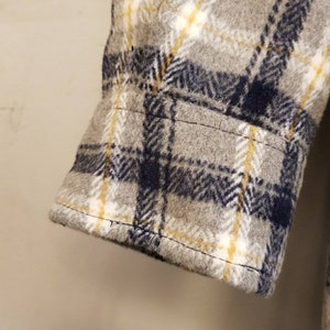May include: A long sleeve shirt cuff featuring a gray, navy blue, and yellow plaid pattern. The fabric has a soft, brushed texture with visible stitching along the hem of the cuff and the sleeve seam, showcasing a classic woven check design.