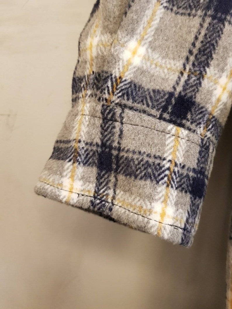 May include: A long sleeve shirt cuff featuring a gray, navy blue, and yellow plaid pattern. The fabric has a soft, brushed texture with visible stitching along the hem of the cuff and the sleeve seam, showcasing a classic woven check design.