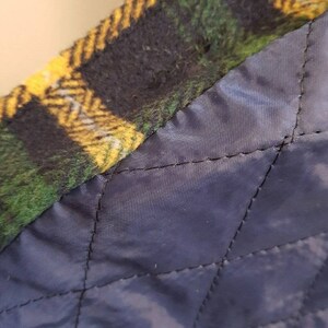 May include: A garment featuring a green, yellow, and black plaid wool-like outer layer joined to a dark navy blue quilted lining. The interior lining displays a diamond-shaped stitched pattern, while the outer edge shows a thick, textured fabric.
