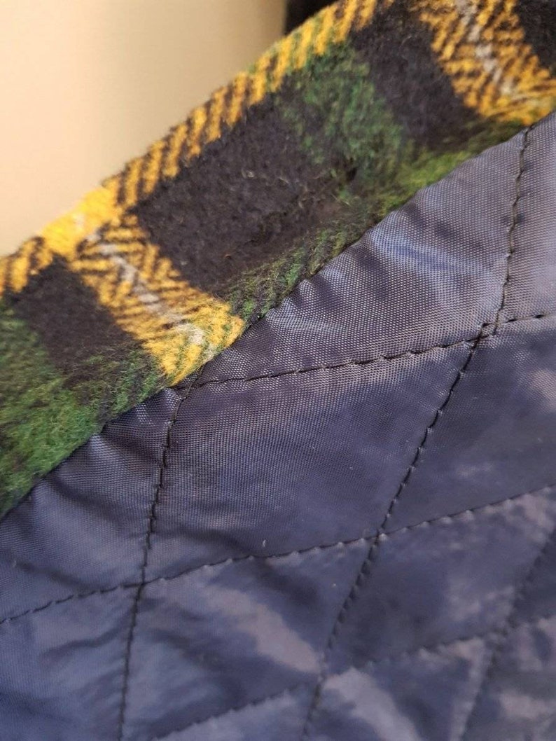 May include: A garment featuring a green, yellow, and black plaid wool-like outer layer joined to a dark navy blue quilted lining. The interior lining displays a diamond-shaped stitched pattern, while the outer edge shows a thick, textured fabric.