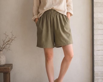 May include: A beige long-sleeved crew neck top paired with olive green elastic-waist shorts featuring side pockets. The outfit is completed with beige canvas lace-up sneakers. The top is tucked into the shorts, which reach mid-thigh length.