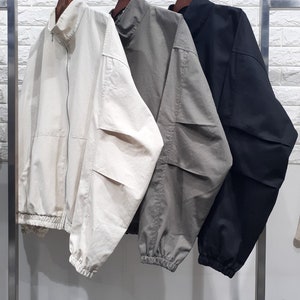 Op de afbeelding: Drie jassen hangen aan een rek. De jas links is crèmekleurig, de middelste jas is grijs en de jas rechts is zwart. Alle drie jassen hebben een staande kraag en een ritssluiting.