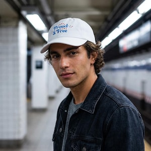 May include: White baseball cap with the text "Proud & Free" in blue script and red stars. The cap is worn by a person wearing a gray shirt and a black denim jacket. The background is a subway station.