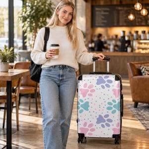May include: A white rolling suitcase featuring a repeating pattern of pink, blue, and purple paw prints with small colorful dots scattered throughout. The luggage has a retractable handle, black corner protectors, and four wheels.