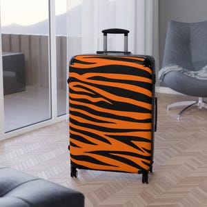 May include: A hard-shell rolling suitcase featuring a vibrant orange and black tiger stripe pattern. The luggage includes a retractable black handle, four multidirectional spinner wheels, and black corner protectors on the top edges.