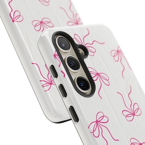 May include: White phone case with a pink bow pattern. The case has a black camera housing and buttons. The bows are drawn with thin lines and are evenly spaced across the case.