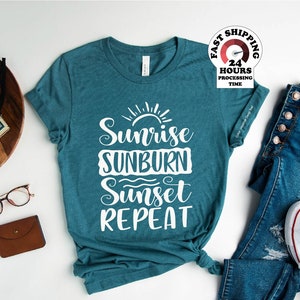 May include: Teal blue t-shirt with white text that reads "Sunrise Sunburn Sunset Repeat". The shirt is laid flat on a white surface with a pair of blue jeans and white sneakers.