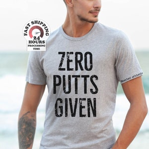 May include: A heather grey t-shirt with the text "ZERO PUTTS GIVEN" printed in black. The text has a distressed look. The t-shirt is being worn by a person standing on a beach.