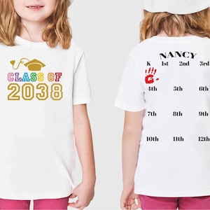 K&ouml;nnte beinhalten: Wei&szlig;es T-Shirt mit einem Regenbogen-Design "CLASS OF 2038" auf der Vorderseite und einem Handabdruck sowie den Klassenstufen von der Vorschule bis zur 12. Klasse auf der R&uuml;ckseite. Der Text "NANCY" ist &uuml;ber den Klassenstufen gedruckt.
