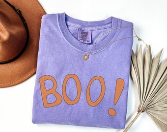 May include: A lavender short-sleeved t-shirt with the word BOO! printed in orange letters across the front. The shirt features a Comfort Colors brand label at the inner neckline and is displayed with a brown hat and a dried palm leaf.