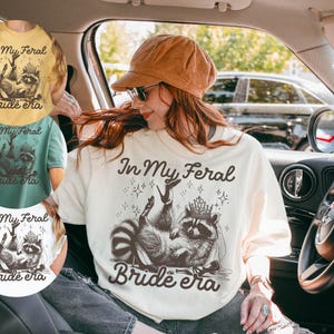 May include: Cream-colored t-shirt with a raccoon graphic wearing a crown and the text "In My Feral Bride Era". The design is in black and white, with stars and a playful font. The shirt is visible in multiple colors.