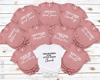 May include: A collection of nine short-sleeved t-shirts arranged in a grid. Eight shirts are dusty rose with white text, and one center shirt is white with black text. Each shirt features unique country music-themed phrases in various fonts.