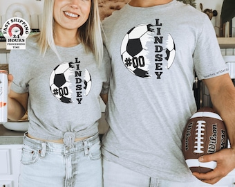 May include: Two heather gray t-shirts featuring a graphic of a soccer ball split by the name LINDSEY and the number #00 in black text. The shirts have short sleeves and a crew neckline. One shirt is tied at the waist, and the other is untied.