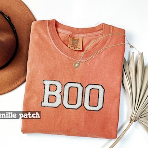 May include: A terracotta-colored short-sleeve t-shirt featuring the word BOO in white chenille patches with black borders. The shirt has a crew neckline and a Comfort Colors brand label at the inner collar. The fabric has a washed, textured appearance.