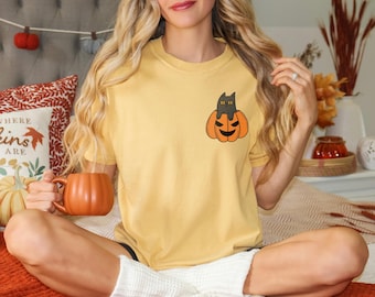 May include: A mustard yellow short-sleeve t-shirt featuring a small graphic on the left chest of a black cat sitting inside an orange jack-o'-lantern pumpkin. The shirt has a crew neckline and a relaxed fit, showcasing a simple seasonal design.