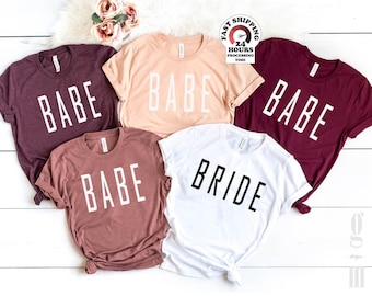 May include: A collection of five short-sleeved crew neck t-shirts in various colors including maroon, peach, mauve, and white. Four shirts feature the word BABE in white or black block lettering, while one white shirt features the word BRIDE.