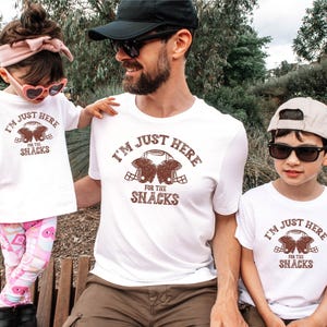 May include: A family wearing white t-shirts with a brown football helmet graphic and the text "I'm just here for the snacks".
