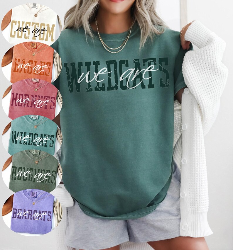 May include: A dark green short-sleeve t-shirt featuring the word WILDCATS in a distressed dark green font with the words we are written in a white cursive script across the center. The shirt is shown in a collection of various colors.