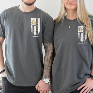 May include: A charcoal grey short-sleeve t-shirt featuring a vertical graphic on the left chest. The graphic depicts a distressed American flag with a gold headset icon and the text DISPATCHER, followed by a personalized name below it.