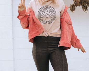 Puede incluir: Una camiseta rosa claro con un gráfico circular de un pavo rodeado por las palabras Happy, Thanksgiving, Grateful, Gather, Family y Love. Se combina con una chaqueta de pana color coral, leggings negros y un gorro rosa claro.