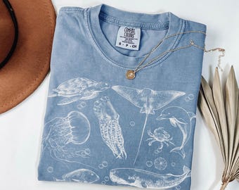 May include: A blue short-sleeve t-shirt featuring a white graphic print of various marine life including a sea turtle, jellyfish, squid, stingray, dolphin, crab, and whale. The shirt has a Comfort Colors brand label at the neck with size S-P-CH.