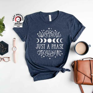 May include: A dark blue heathered t-shirt featuring a white graphic print of moon phases, floral elements, and stars, with the text "JUST A PHASE" centered in the design. The shirt is displayed with a brown handbag, a watch, glasses, and a small plant.