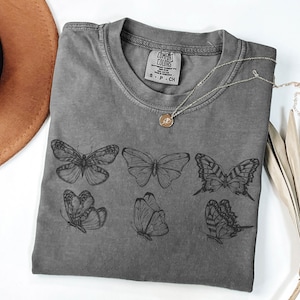 Puede incluir: Una camiseta de manga corta gris carb&oacute;n con un estampado gr&aacute;fico en tinta negra de seis mariposas diferentes. La camiseta tiene un cuello redondo y una etiqueta de la marca Comfort Colors impresa en el interior del cuello con la talla S-P-CH.