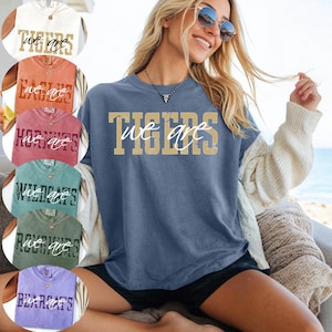 May include: A blue short-sleeve t-shirt featuring the text "WE ARE" in white script layered over the word "TIGERS" in large, distressed gold block letters. The shirt has a crew neckline and a relaxed fit, shown alongside other color variations.