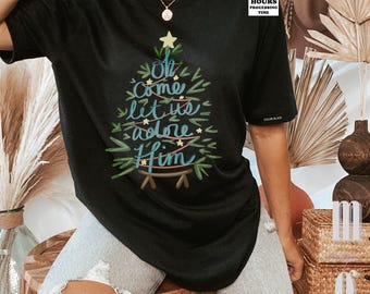 May include: A black short-sleeved t-shirt featuring a graphic of a stylized Christmas tree formed by green brushstrokes and a yellow star. The text "Oh come let us adore Him" is written in blue cursive script across the center of the tree.