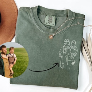 May include: An olive green t-shirt featuring a white line art illustration of an adult holding a child. The shirt includes a Comfort Colors brand tag at the neck and a circular inset photograph showing the same adult and child in a field.