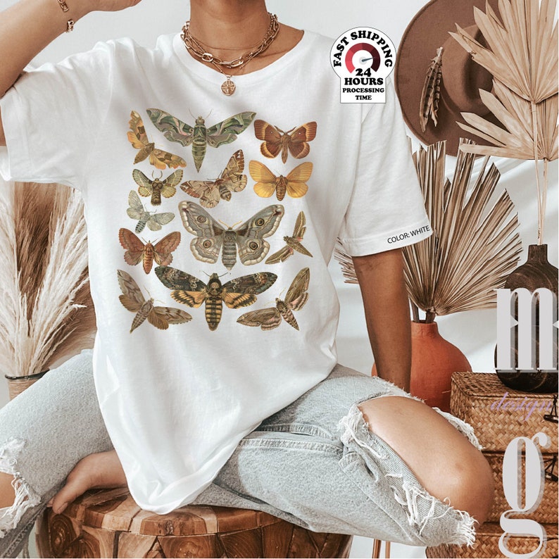 May include: White t-shirt with a print of various butterflies and moths in shades of brown, yellow, and green. The text "COLOR: WHITE" is visible on the shirt's sleeve.