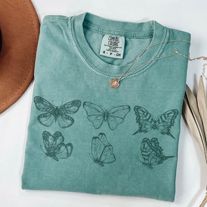 Puede incluir: Una camiseta de manga corta color verde salvia con un estampado gr&aacute;fico de seis mariposas distintas en tinta verde oscuro. La prenda tiene cuello redondo y una etiqueta de la marca Comfort Colors en el cuello interior con el texto S - P - CH.