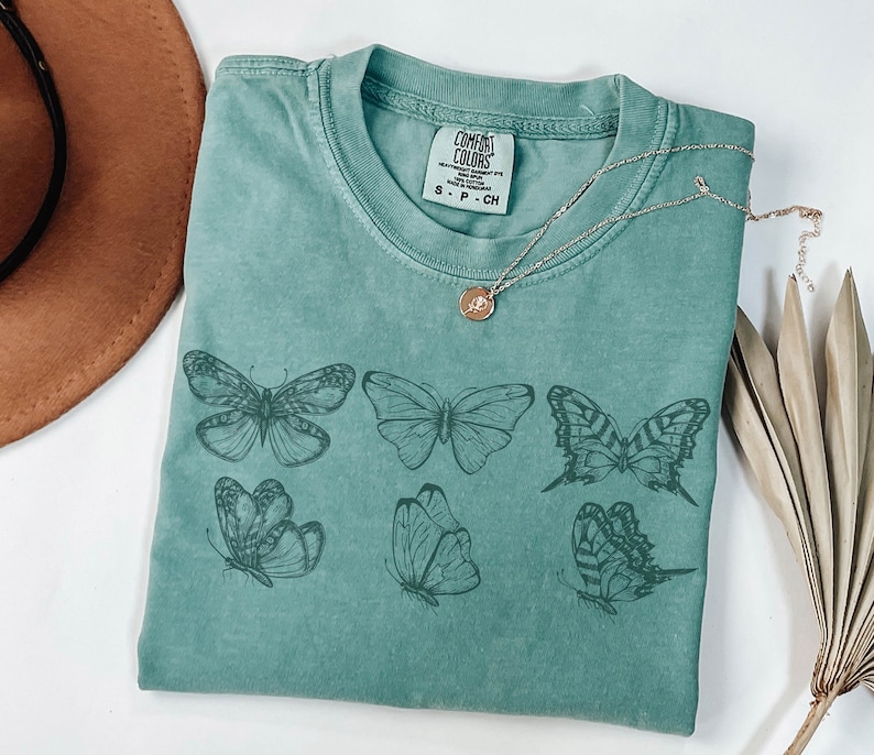 Puede incluir: Una camiseta de manga corta color verde salvia con un estampado gr&aacute;fico de seis mariposas distintas en tinta verde oscuro. La prenda tiene cuello redondo y una etiqueta de la marca Comfort Colors en el cuello interior con el texto S - P - CH.