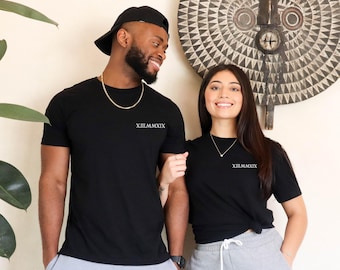 May include: Two black short-sleeved t-shirts featuring the Roman numeral text XII.MMXIX printed in white on the upper left chest. The shirts are paired with light gray sweatpants, a gold chain necklace, and a black baseball cap worn backwards.