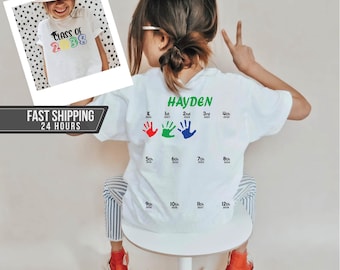 May include: A white t-shirt featuring the name Hayden in green text, followed by a grid of school years from K to 12th grade. The design includes three colorful handprints in red, green, and blue, and a small inset photo of a child wearing the shirt.