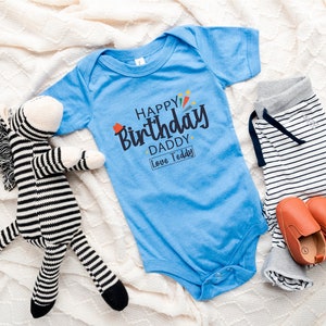 May include: A blue baby onesie with the text "Happy Birthday Daddy Love Teddy" in black and colorful lettering. A striped zebra stuffed animal and a pair of brown baby shoes are also in the image.