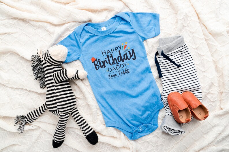 May include: A blue baby onesie with the text "Happy Birthday Daddy Love Teddy" in black and colorful lettering. A striped zebra stuffed animal and a pair of brown baby shoes are also in the image.
