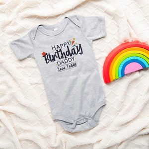 May include: A gray baby onesie with the text "Happy Birthday Daddy Love Teddy" in black. A rainbow-colored wooden toy is in the background.