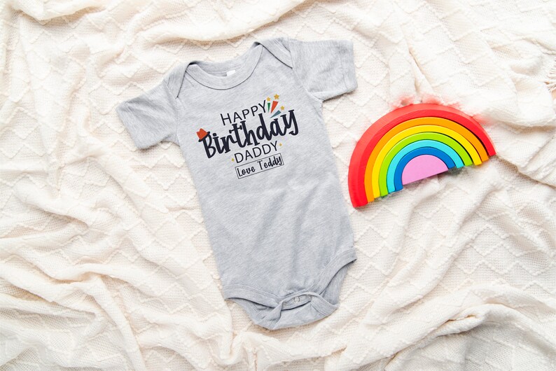 May include: A gray baby onesie with the text "Happy Birthday Daddy Love Teddy" in black. A rainbow-colored wooden toy is in the background.