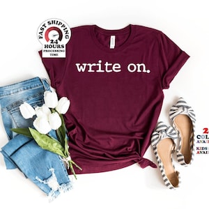 May include: A maroon short-sleeved t-shirt with the white text "write on." printed on the chest. The shirt is displayed alongside a pair of blue denim jeans, a bouquet of white tulips, and a pair of black and white striped pointed-toe flats.