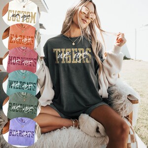 May include: A charcoal gray short-sleeve t-shirt featuring the gold text TIGERS and the white cursive text we are. The shirt has a crew neckline and a relaxed fit. Additional color options are displayed in circular insets on the left side.