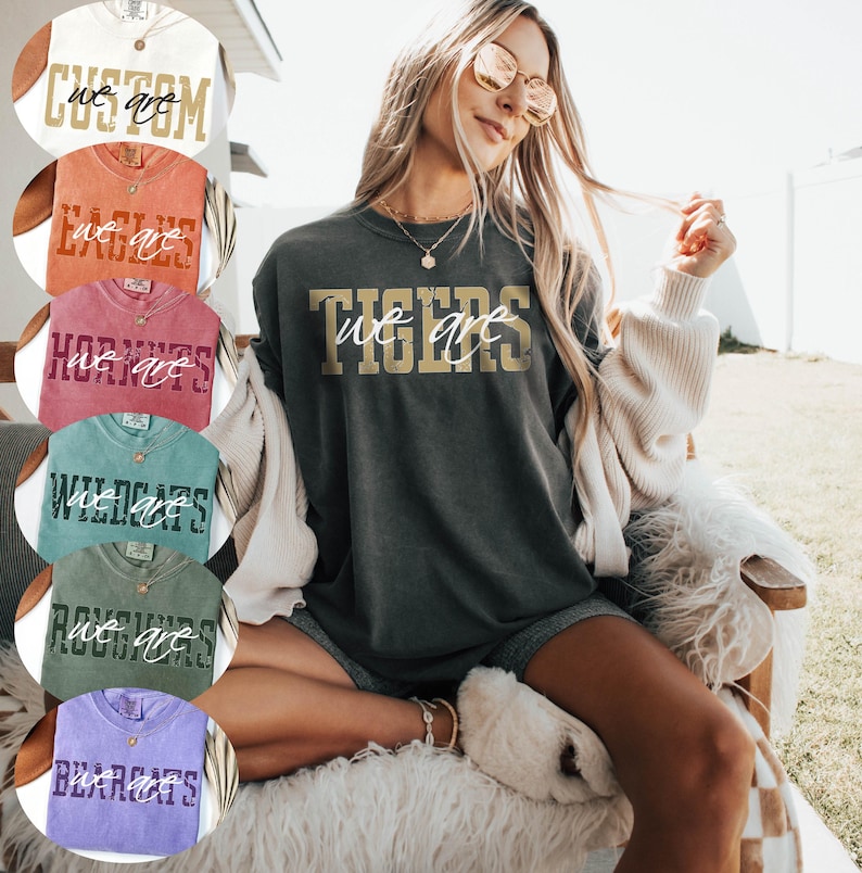 May include: A charcoal gray short-sleeve t-shirt featuring the gold text TIGERS and the white cursive text we are. The shirt has a crew neckline and a relaxed fit. Additional color options are displayed in circular insets on the left side.