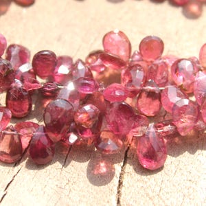 May include: A strand of pink gemstone beads, each bead is faceted and shaped like a teardrop. The beads are strung together on a clear thread.
