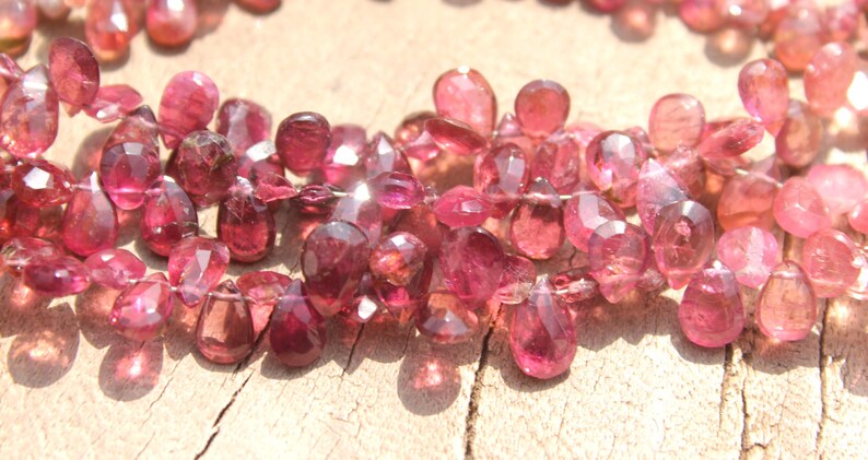 May include: A strand of pink gemstone beads, each bead is faceted and shaped like a teardrop. The beads are strung together on a clear thread.