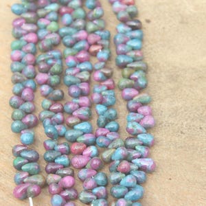 May include: A strand of teardrop-shaped gemstone beads in shades of pink, green, and blue. The beads are strung on a white thread and are arranged in a row.