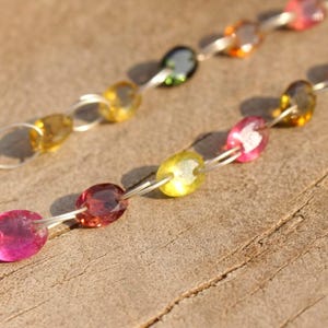 Pode incluir: Um colar de corrente de prata com pedras preciosas ovais em tons de rosa, amarelo e verde. O colar est&aacute; sobre uma superf&iacute;cie de madeira.