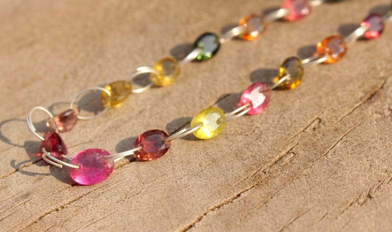 Pode incluir: Um colar de corrente de prata com pedras preciosas ovais em tons de rosa, amarelo e verde. O colar est&aacute; sobre uma superf&iacute;cie de madeira.