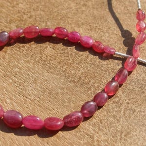 May include: A strand of oval-shaped, deep pink gemstones. The beads are strung on a silver-colored wire, with a pink tassel at the end. The gemstones are a vibrant, saturated color, and the background is a warm, wooden surface.