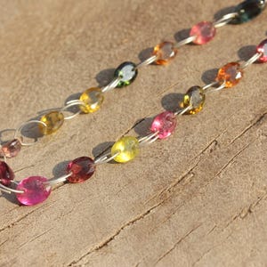 Pode incluir: Um colar de corrente de prata com pedras preciosas ovais de v&aacute;rias cores, incluindo rosa, amarelo, verde e marrom. O colar est&aacute; sobre uma superf&iacute;cie de madeira.