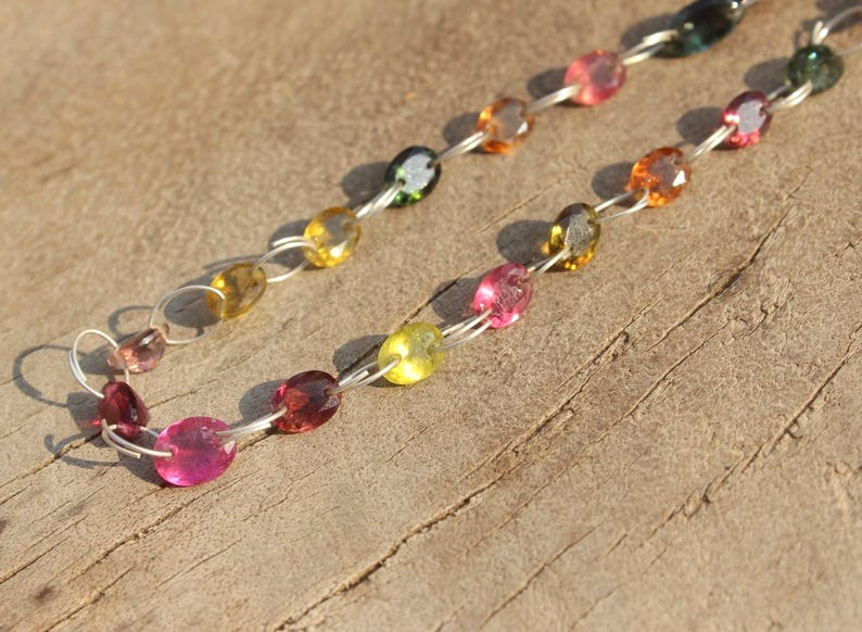 Pode incluir: Um colar de corrente de prata com pedras preciosas ovais de v&aacute;rias cores, incluindo rosa, amarelo, verde e marrom. O colar est&aacute; sobre uma superf&iacute;cie de madeira.